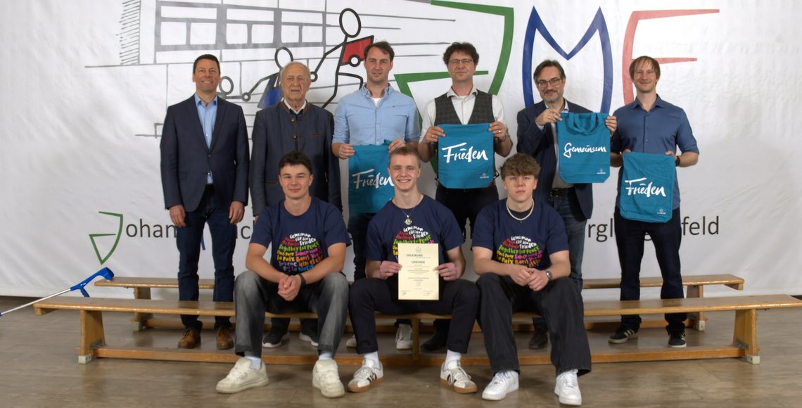 Drei Schüler auf einer Bank, dahinter für fünf Personen, die zum Gewinn des Schülerwettbewerbs gratulieren.