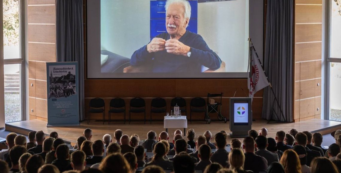 Publikum im Hörsaal blickt auf eine große Leinwandprojektion.