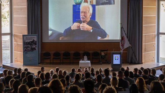 Publikum im Hörsaal blickt auf eine große Leinwandprojektion.
